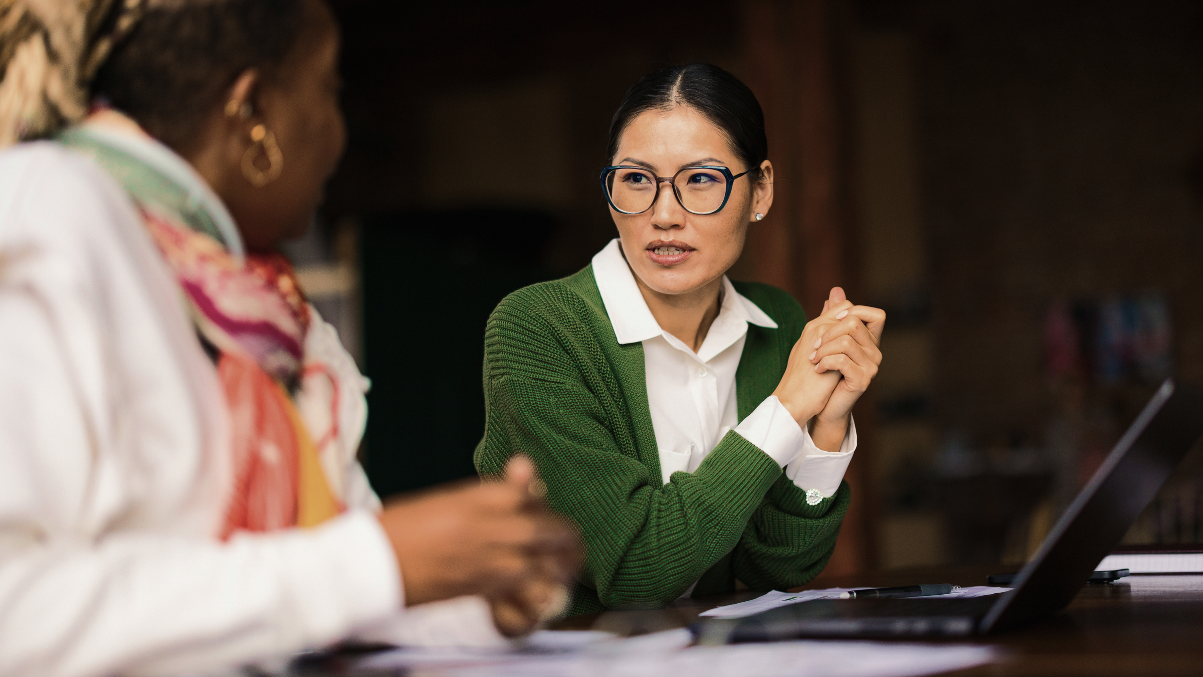 An Asian woman in glasses and a green cardigan looks attentively at another woman across a table with a laptop.