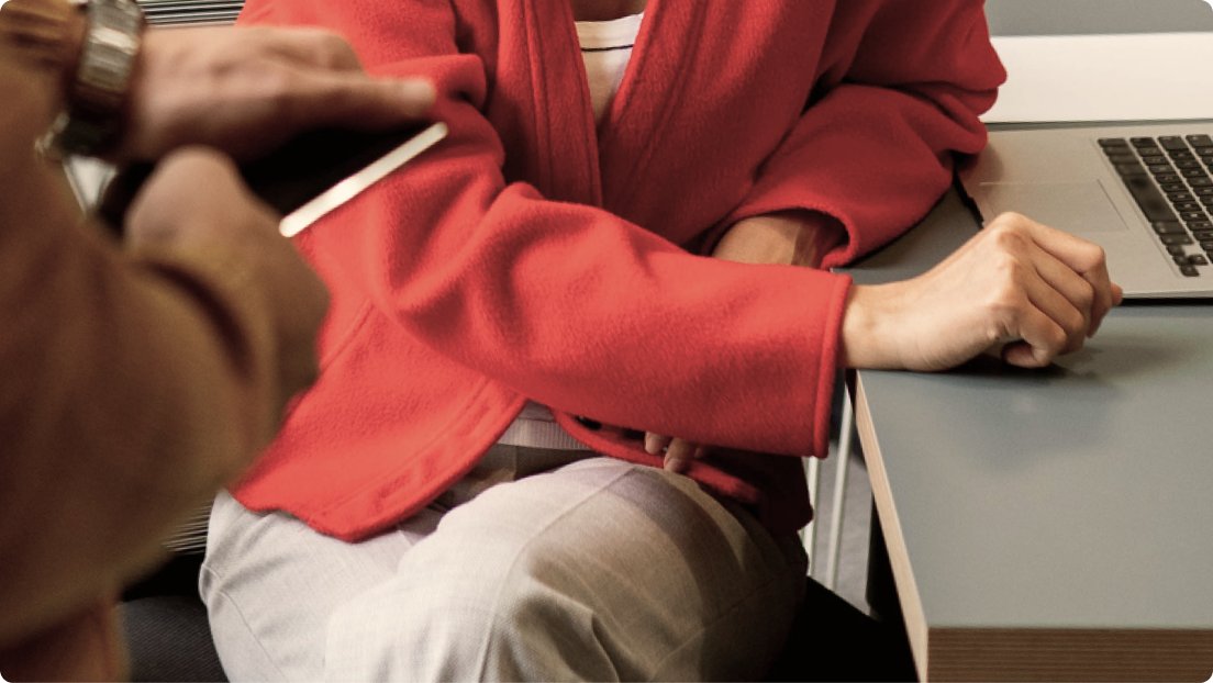 A person in a red jacket with a clenched fist sits at a desk with a laptop, as another person holds a smartphone nearby.