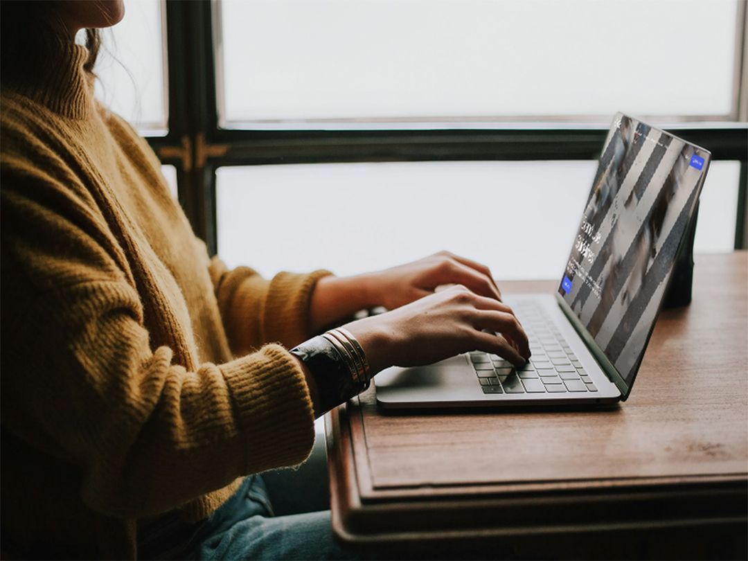 Person in a yellow sweater typing on a laptop on a wooden table.
