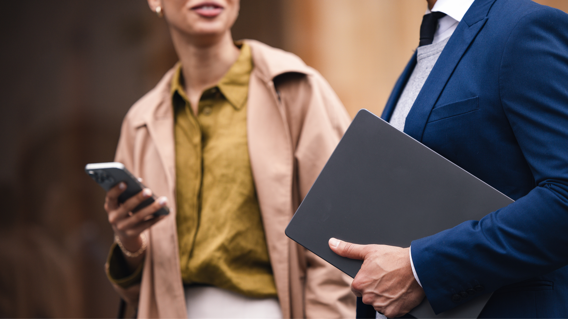 A woman holding a smartphone and a man holding a laptop, dressed in professional attire.