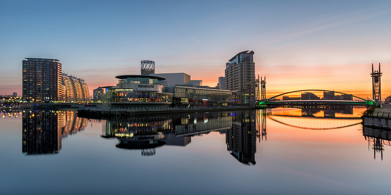 Modern city skyline with buildings and a green-lit bridge reflected in calm water under a colorful sunset sky.