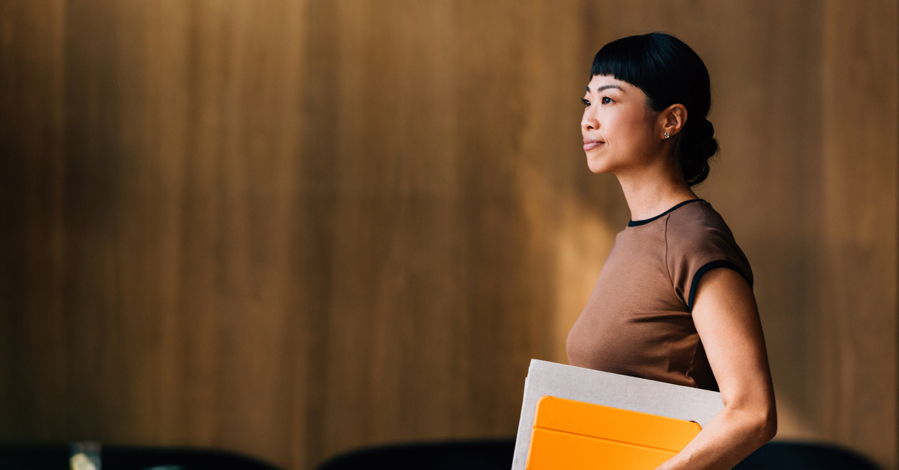 A woman with dark hair and bangs looks left, holding a folder and an orange tablet.