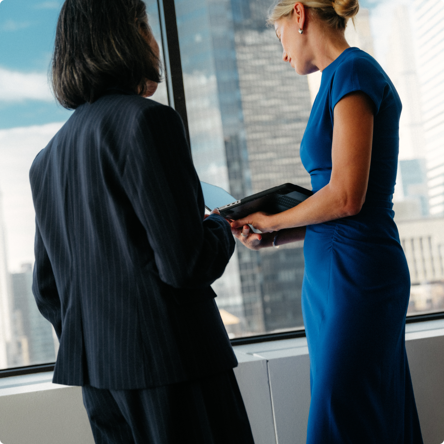 Two businesswomen reviewing tablets by a large office window.