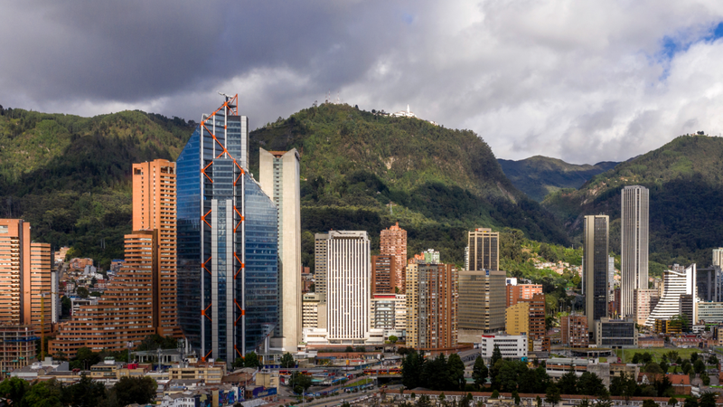 Cityscape with a tall blue skyscraper among other buildings, backed by green mountains and a cloudy sky.