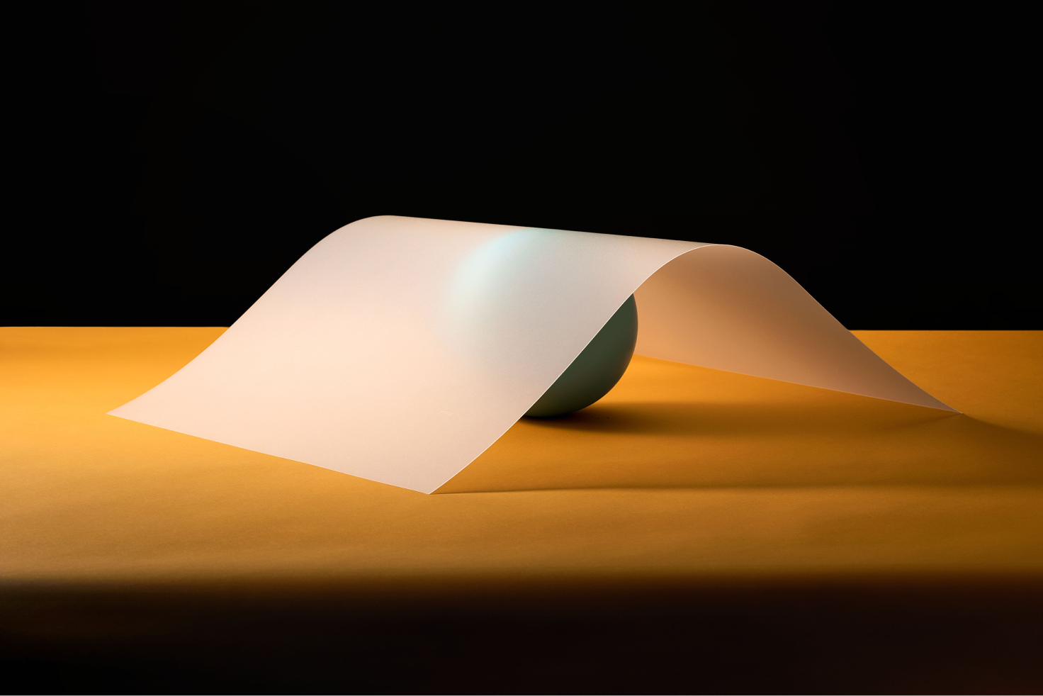 A translucent paper sheet arches over a teal sphere on an orange surface with a black background.