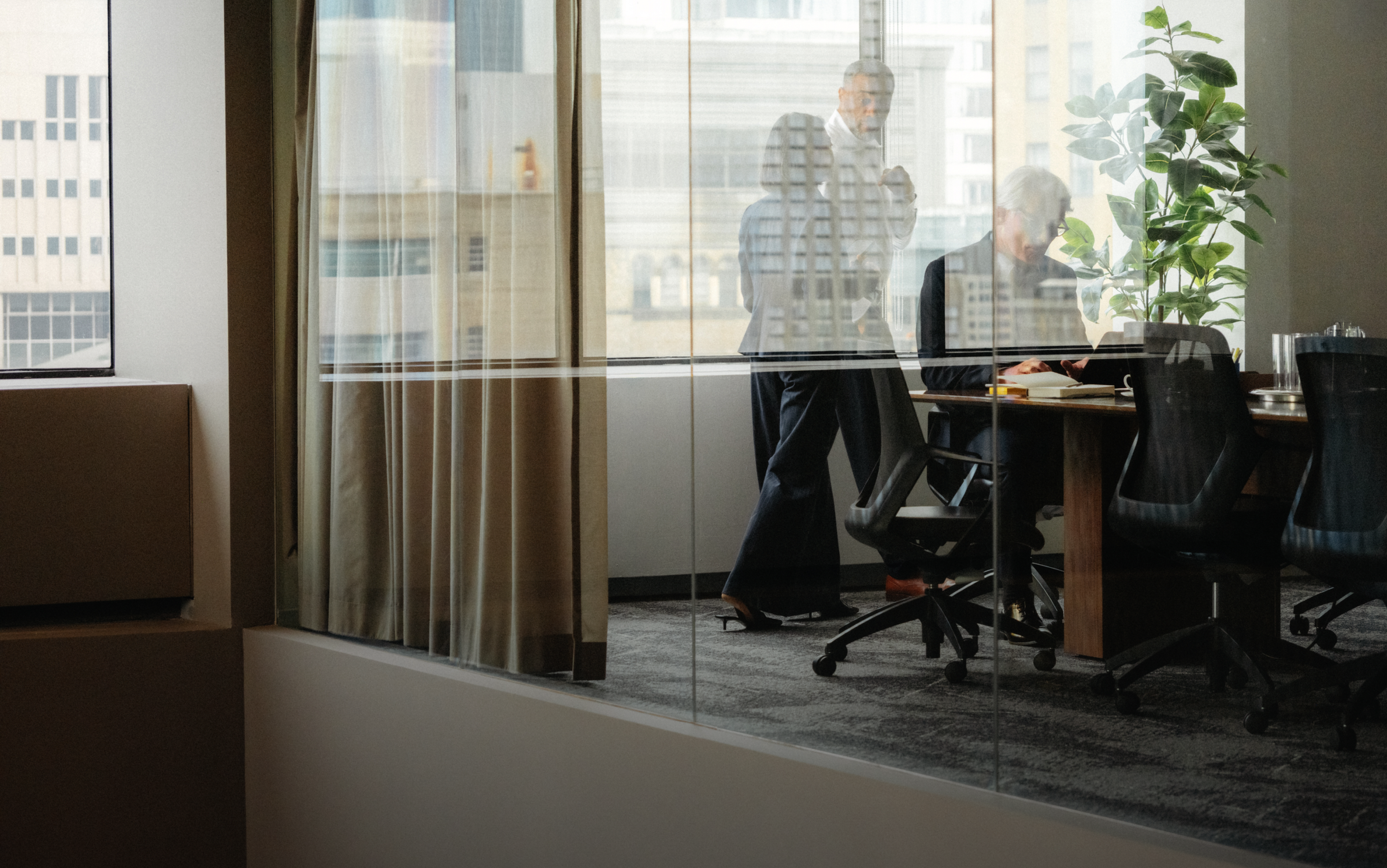 Three people in a modern office meeting room, viewed through a glass partition with city reflections.