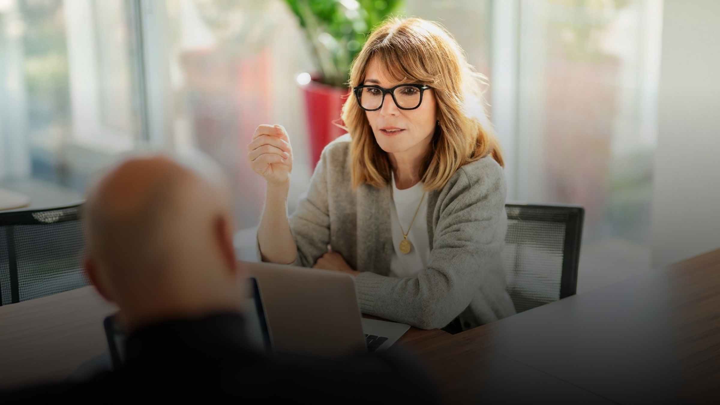 Woman in glasses talks and gestures to a person across a table with a laptop.