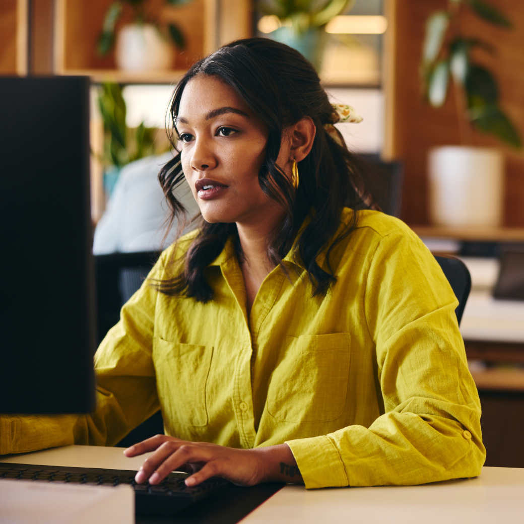 A focused woman in a yellow shirt works on a computer in an office.