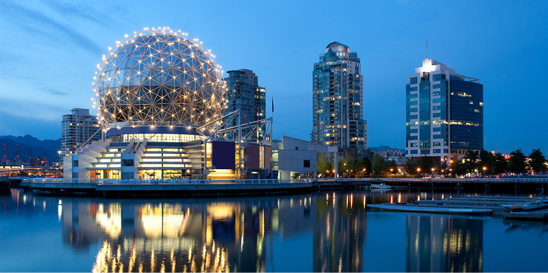 A brightly lit geodesic dome and city skyline reflected on a body of water at dusk.
