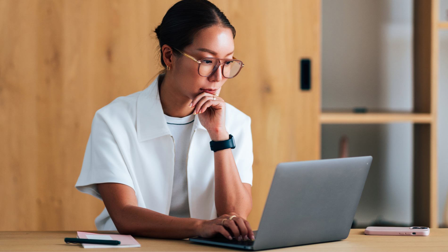 Woman in glasses rests her chin on her hand while reading her laptop at a wooden desk