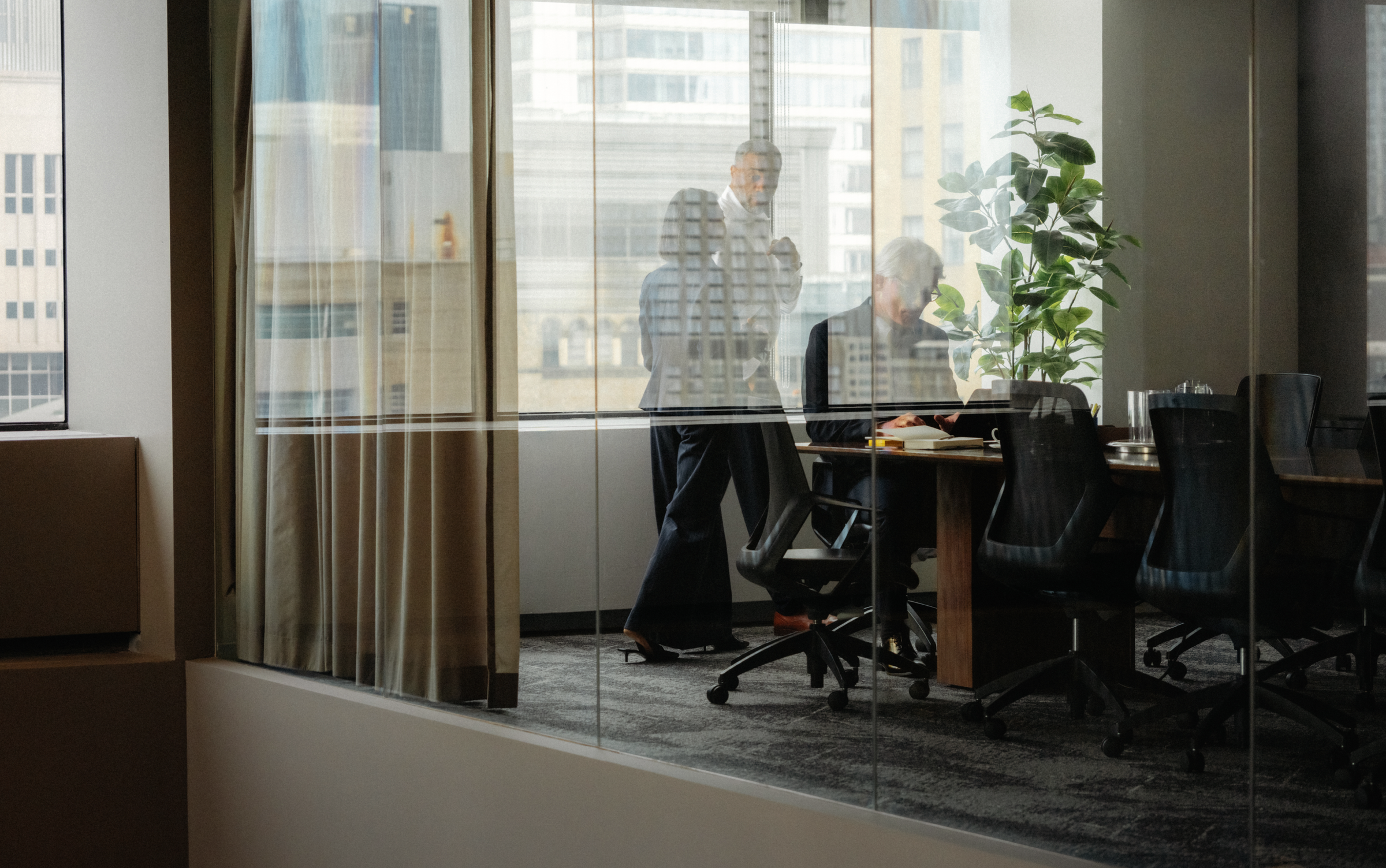 Three professionals in a modern conference room, visible through a glass wall, with a city view outside.