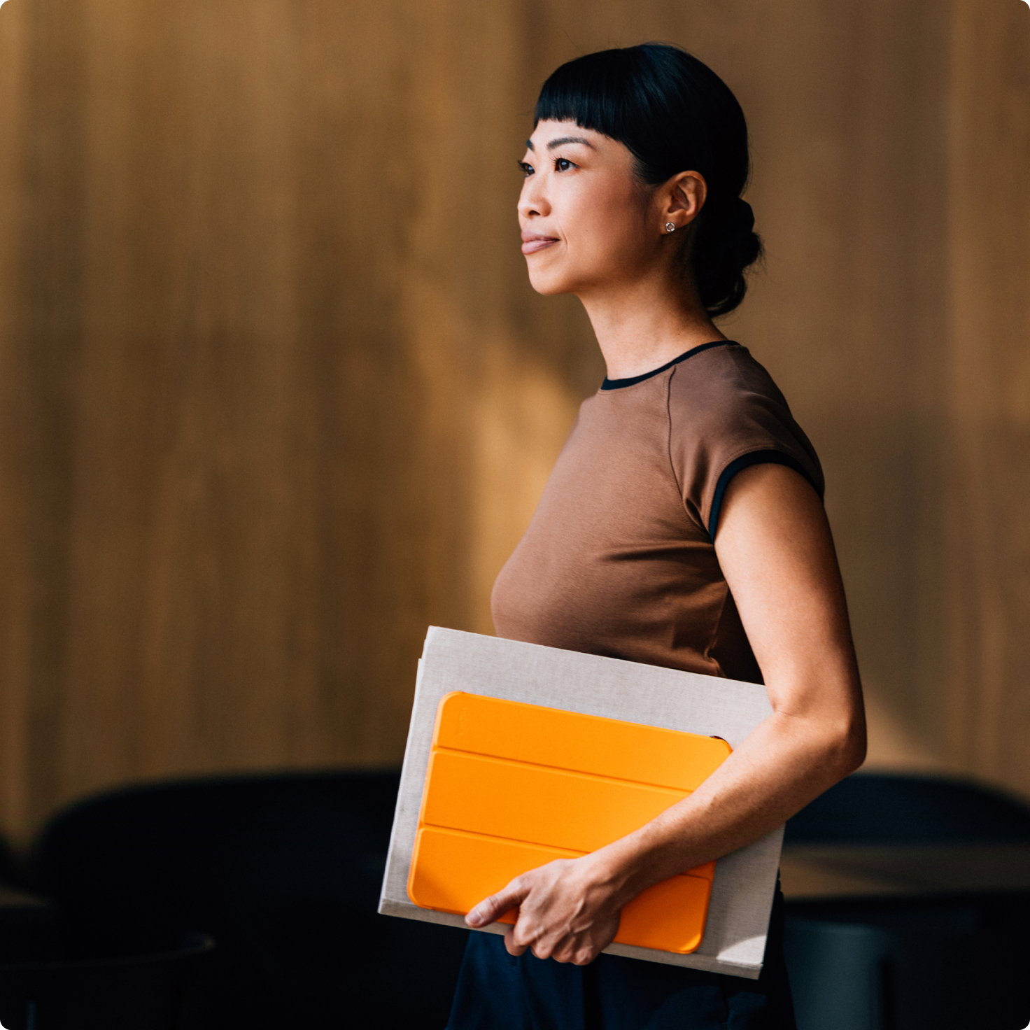 An Asian woman with a bob haircut holds an orange tablet and a gray folder, looking to the right with a slight smile.
