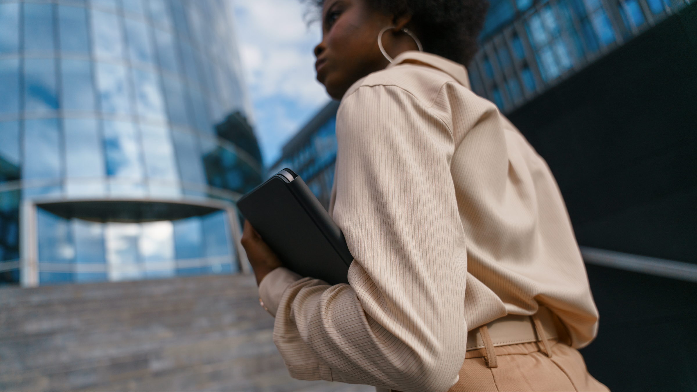 Woman in professional attire holding a tablet outside a modern glass building.