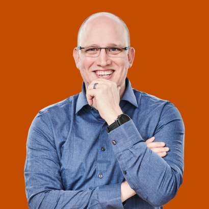 A smiling bald man wearing glasses and a blue shirt, with his hand on his chin.