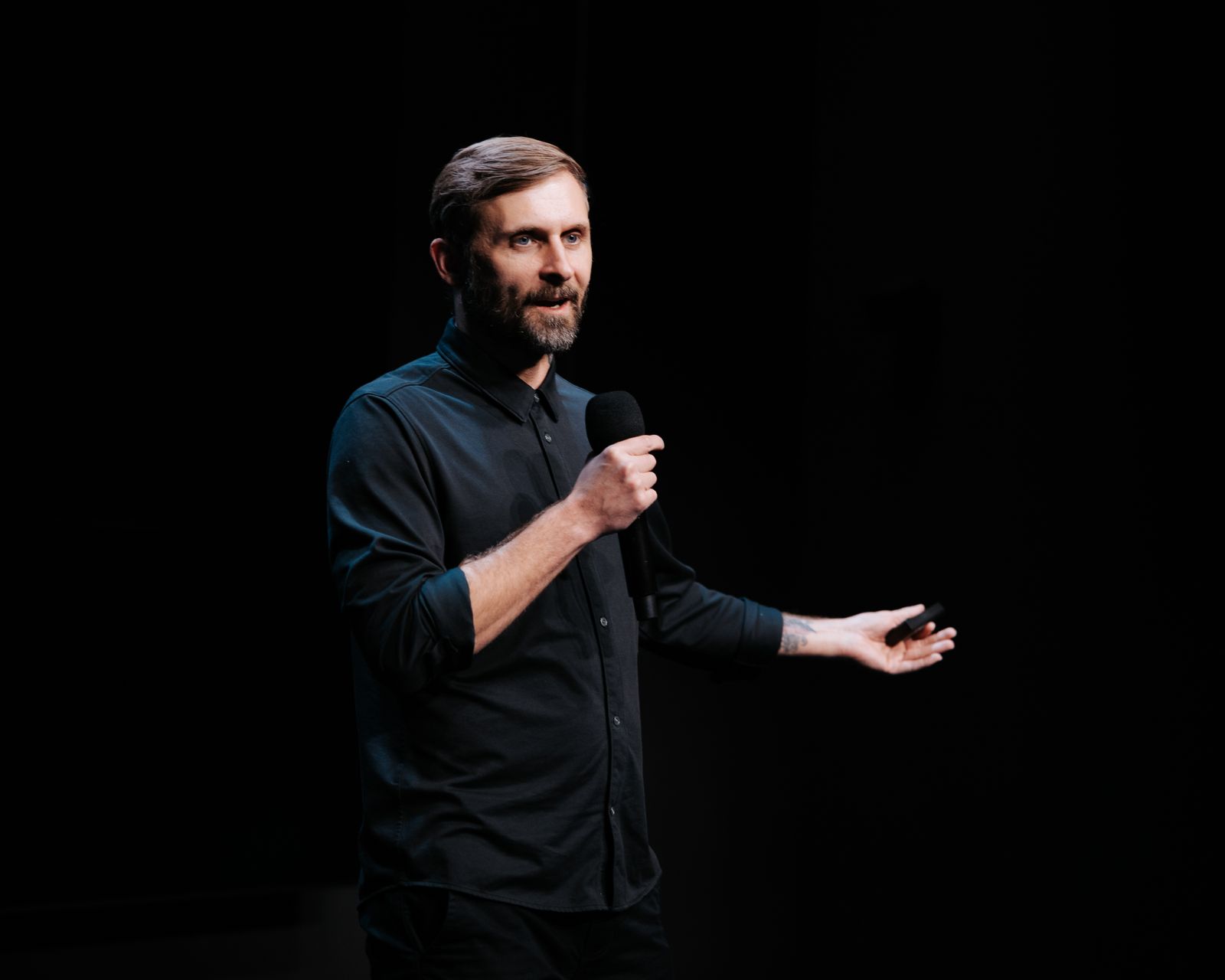 A bearded man in a dark shirt speaks into a microphone, holding a presentation remote in his other hand, against a black background.