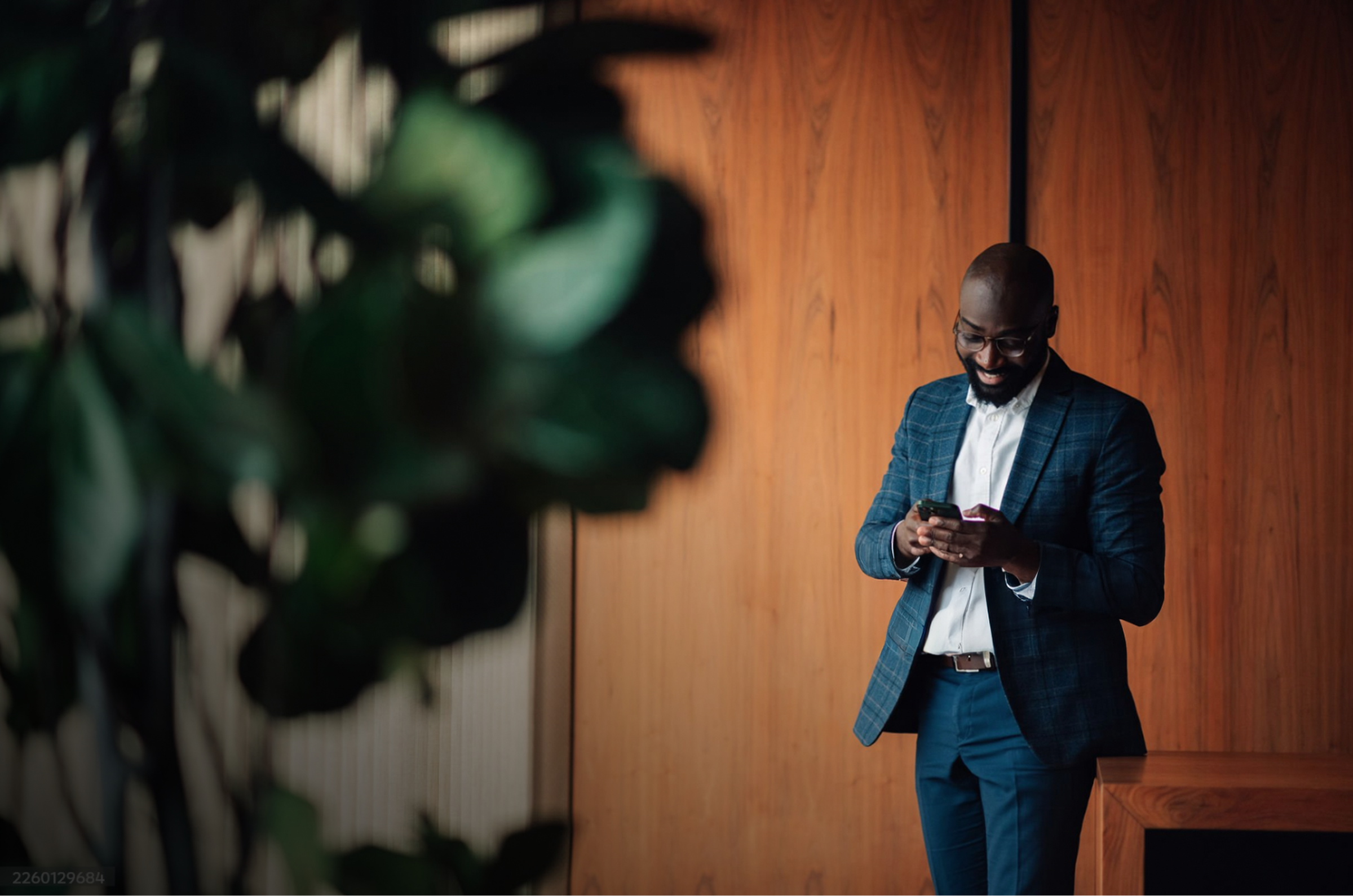 A smiling Black man in a suit and glasses looks at his phone.