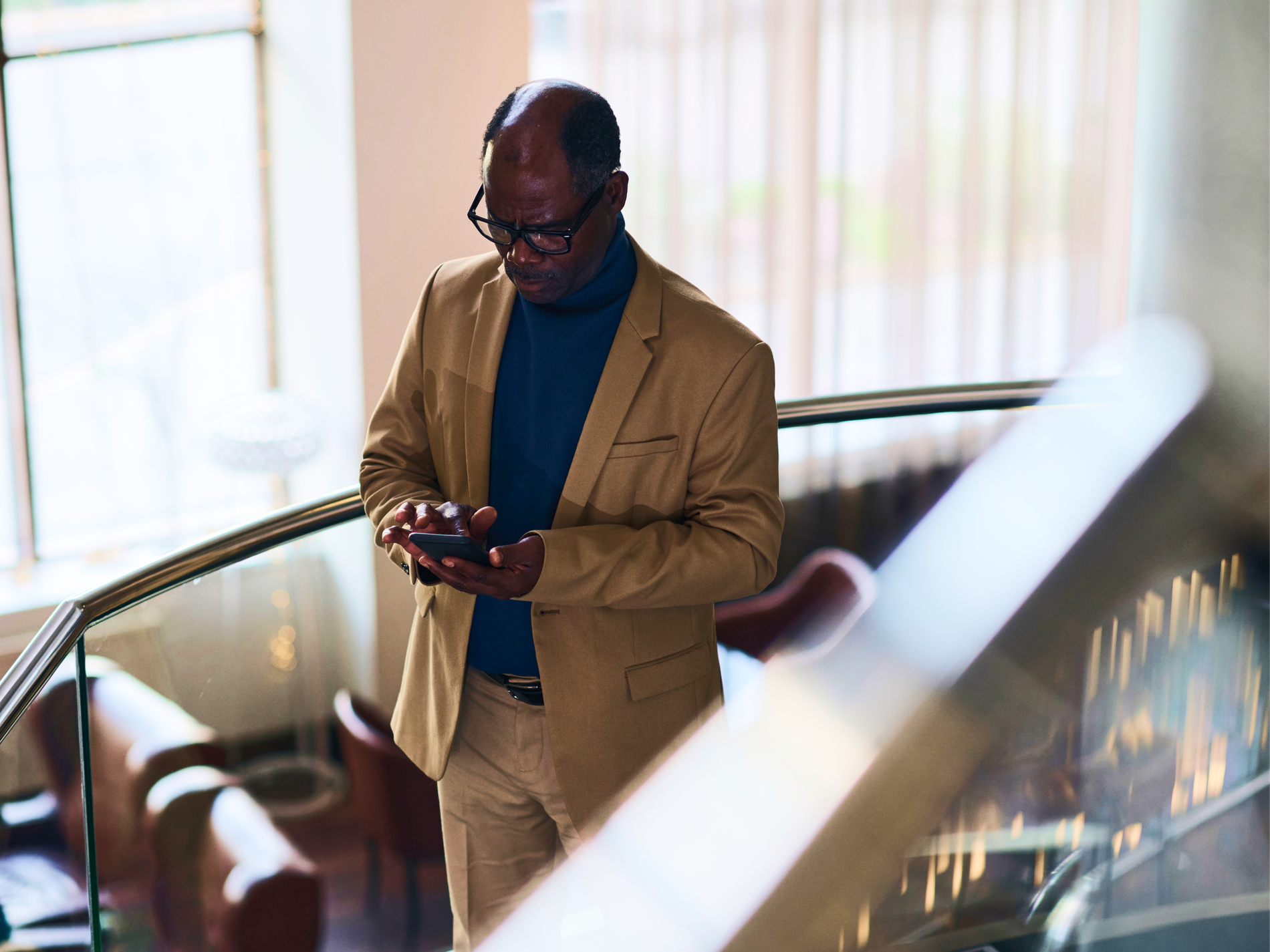 Older Black man in a tan blazer and blue turtleneck using a smartphone on a modern staircase.