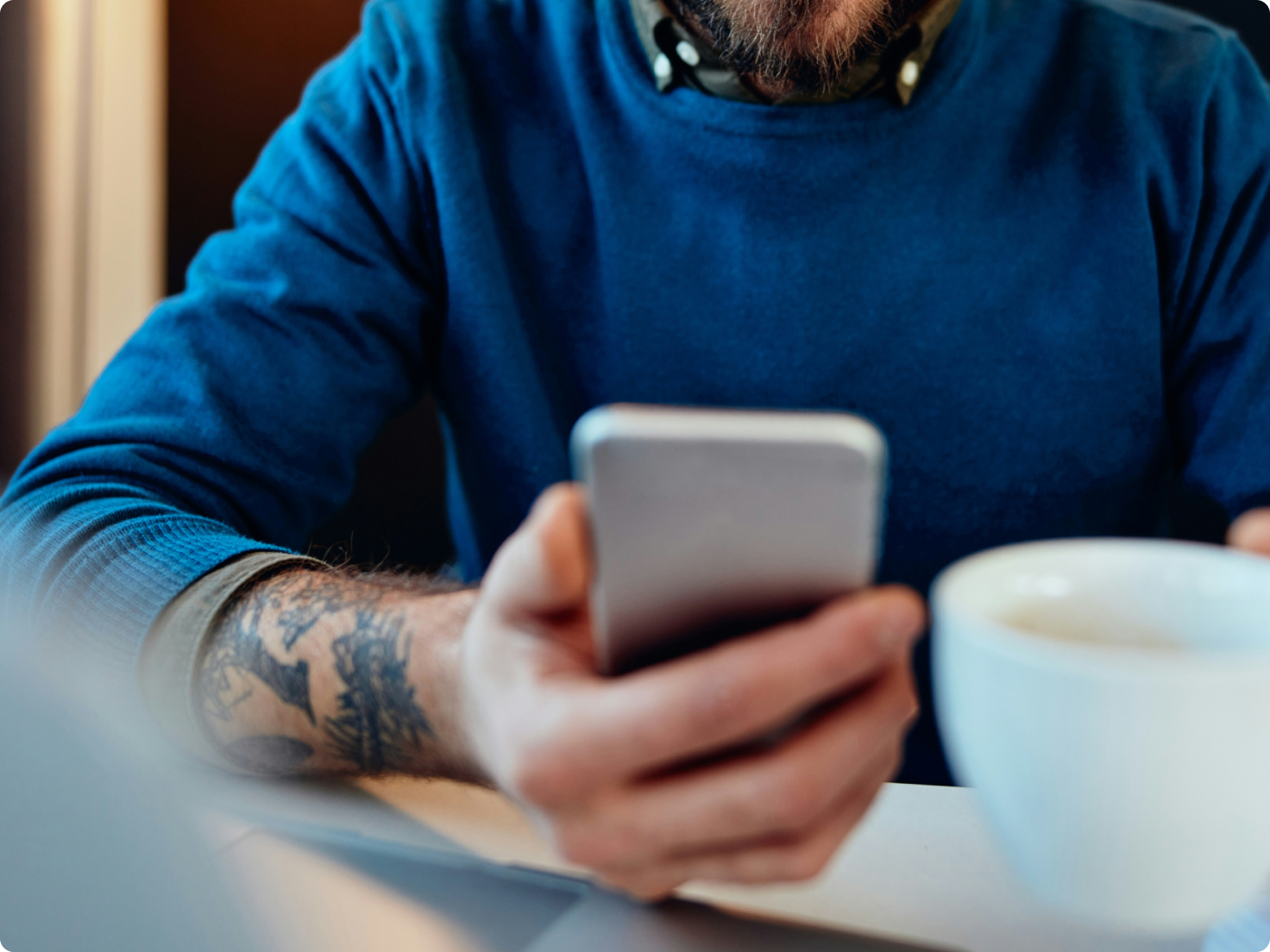 Man in blue sweater with tattooed arm holding a smartphone and a coffee cup.