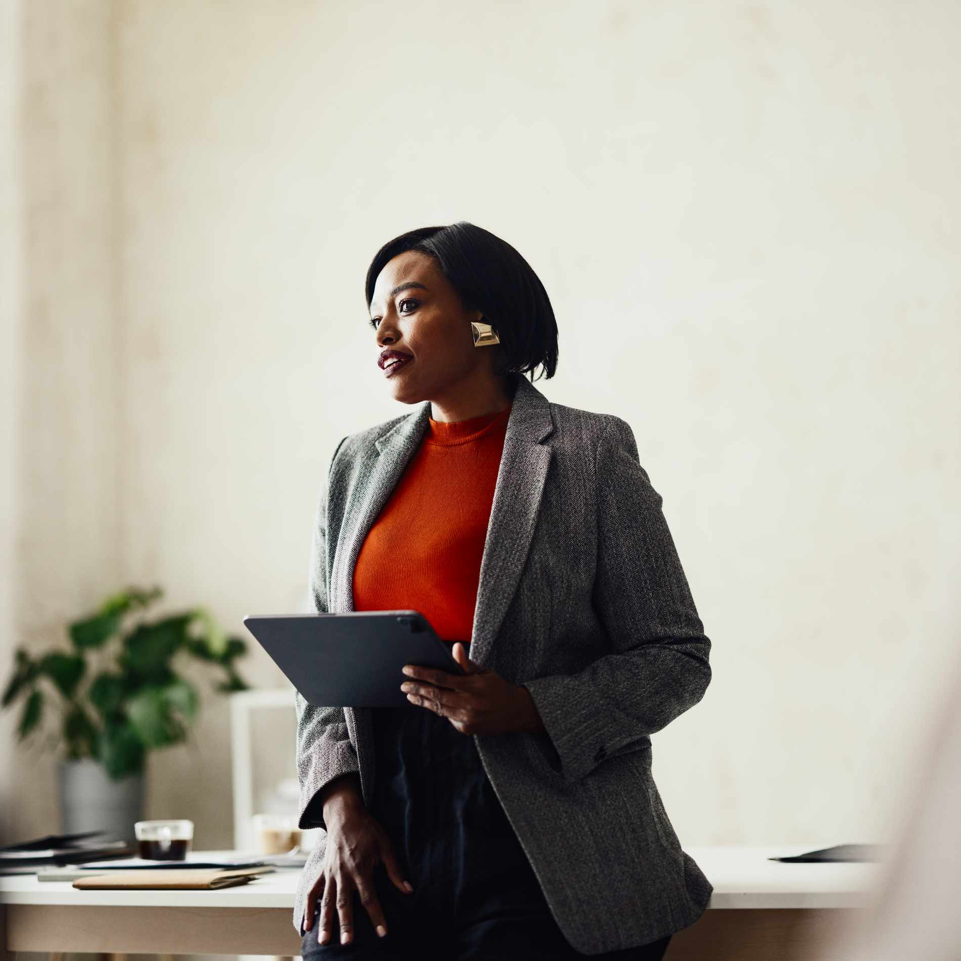 A Black woman in a blazer and orange top holds a tablet while leaning on a desk.