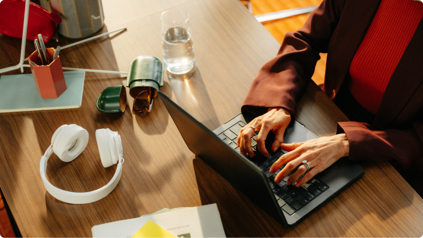 Hands typing on a laptop on a wooden desk with headphones, a water glass, and sunglasses.