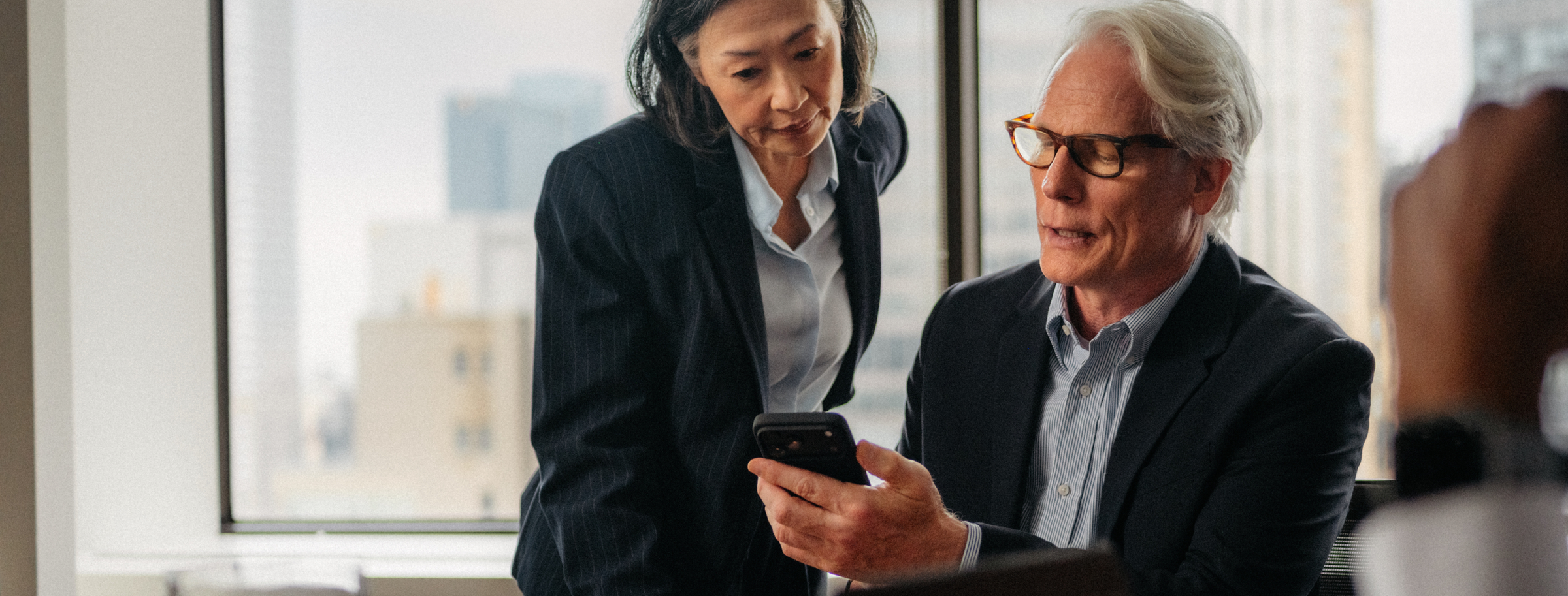 An older man and woman in business attire looking at a smartphone together in an office.