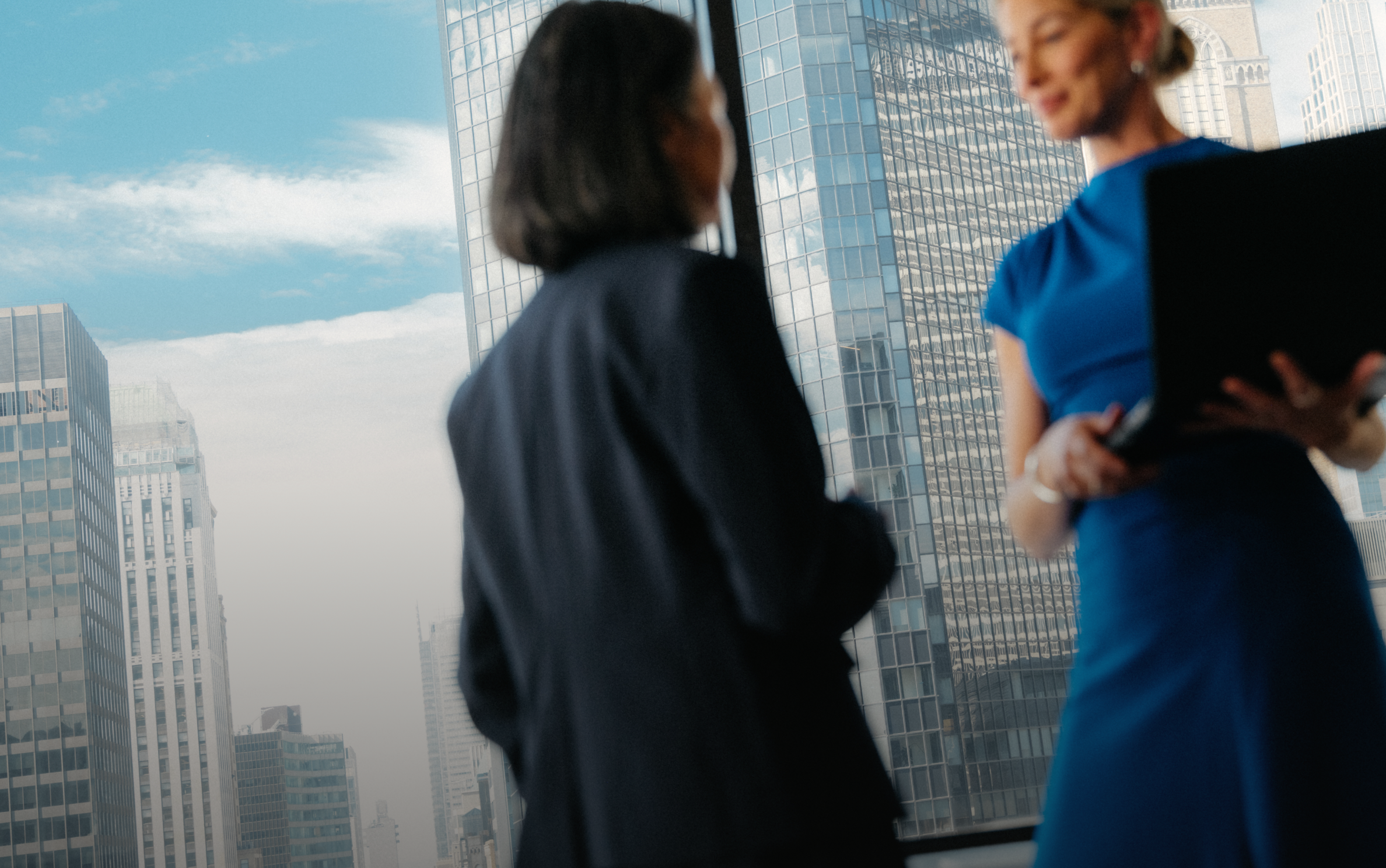 Two businesswomen in an office overlooking a city skyline, one in blue holding a laptop.