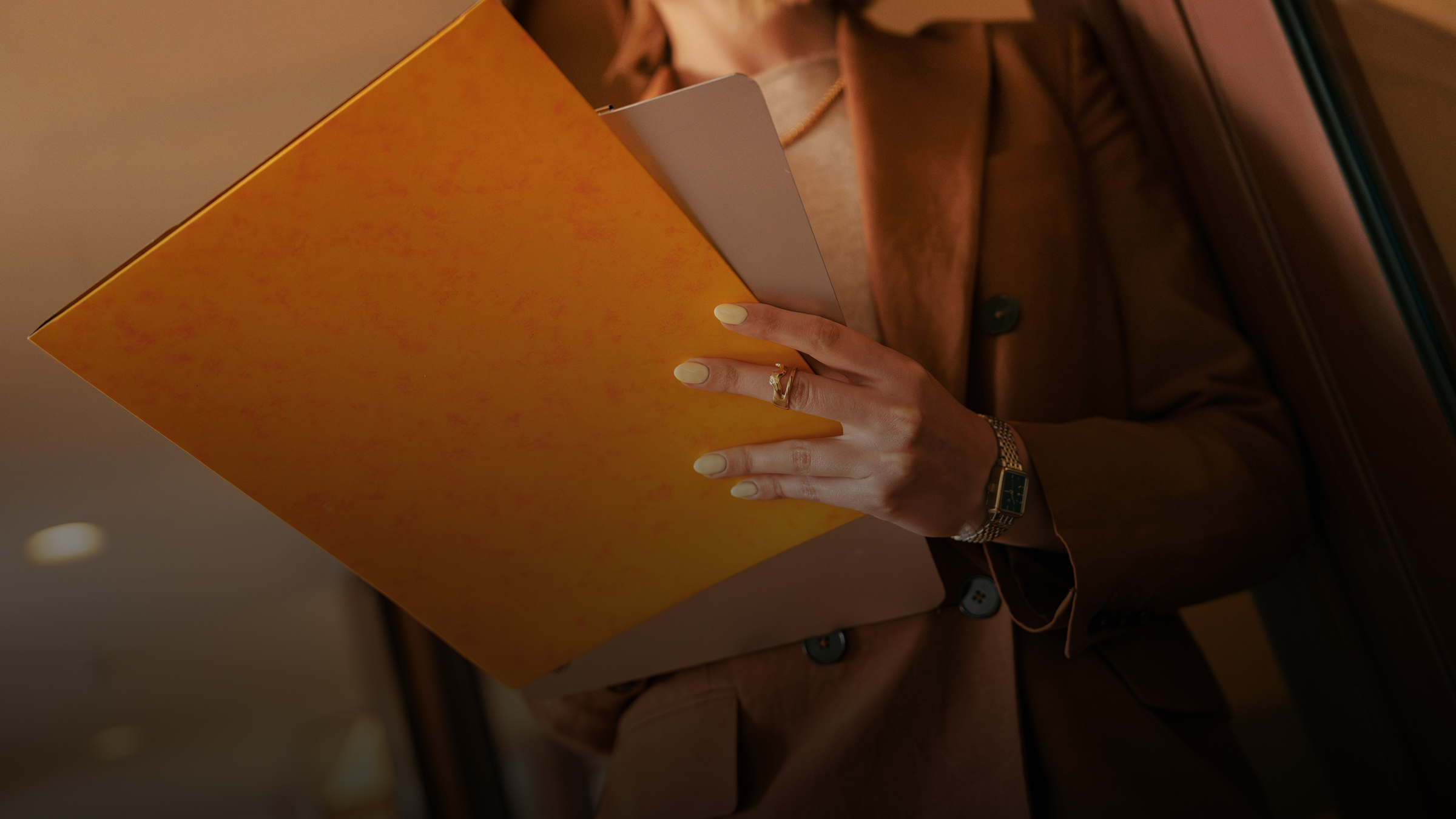 A person in a brown suit holds a bright yellow folder and a light notebook, with a ring and watch on their hand.