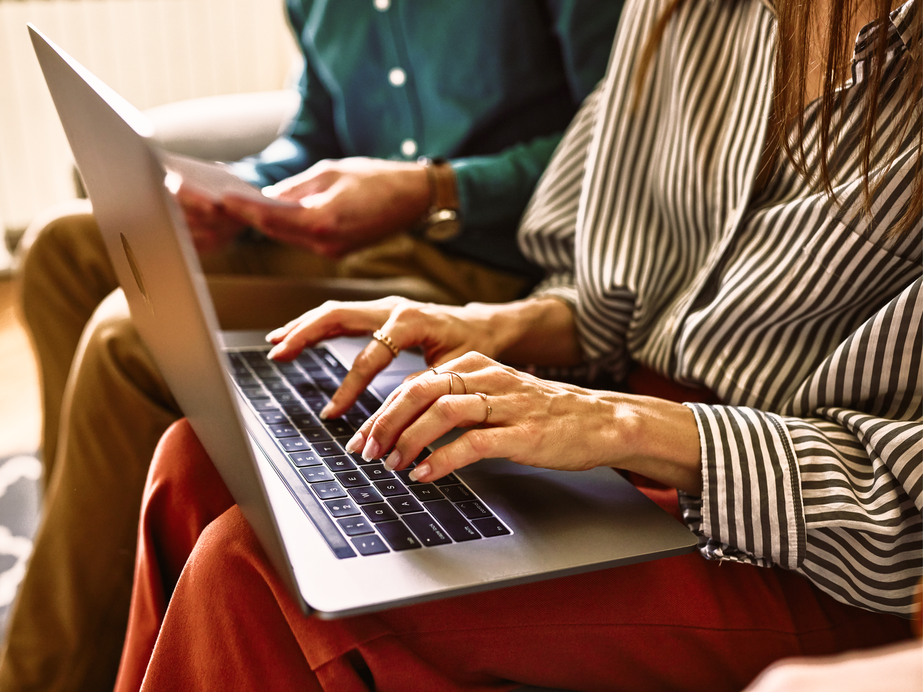 A person's hands type on a silver laptop, with another person holding papers in the blurred background.