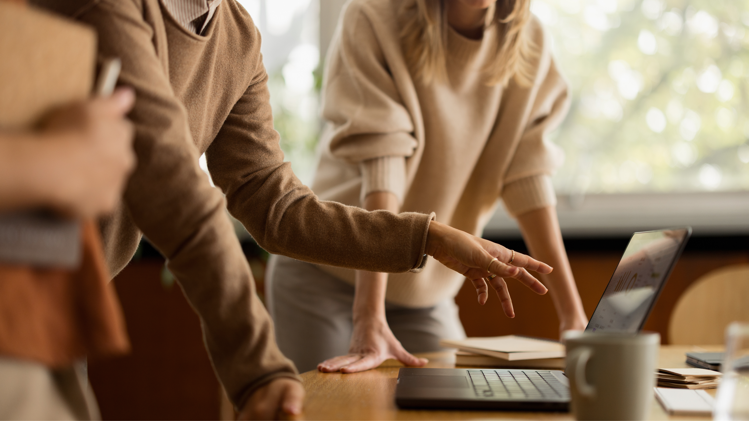 Two colleagues lean over a desk, pointing at a laptop as they work through a document together.