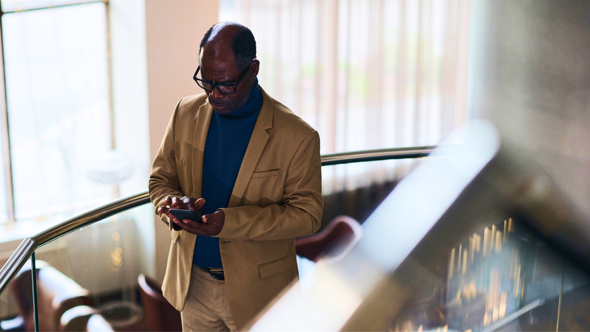 Older Black man in a blazer and turtleneck looking at his smartphone on a staircase.