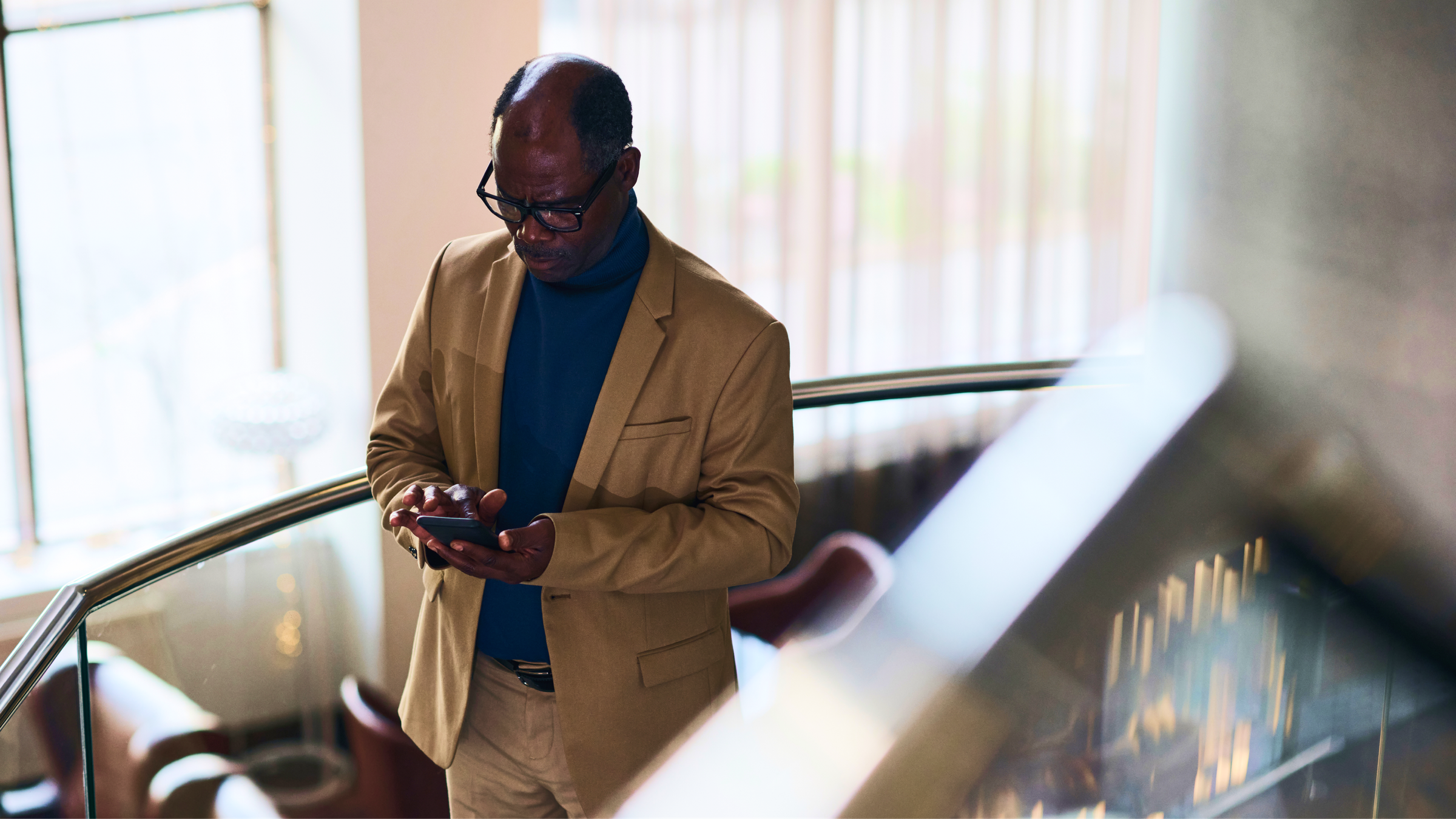 An older Black man in a tan blazer and blue turtleneck looks at his smartphone.
