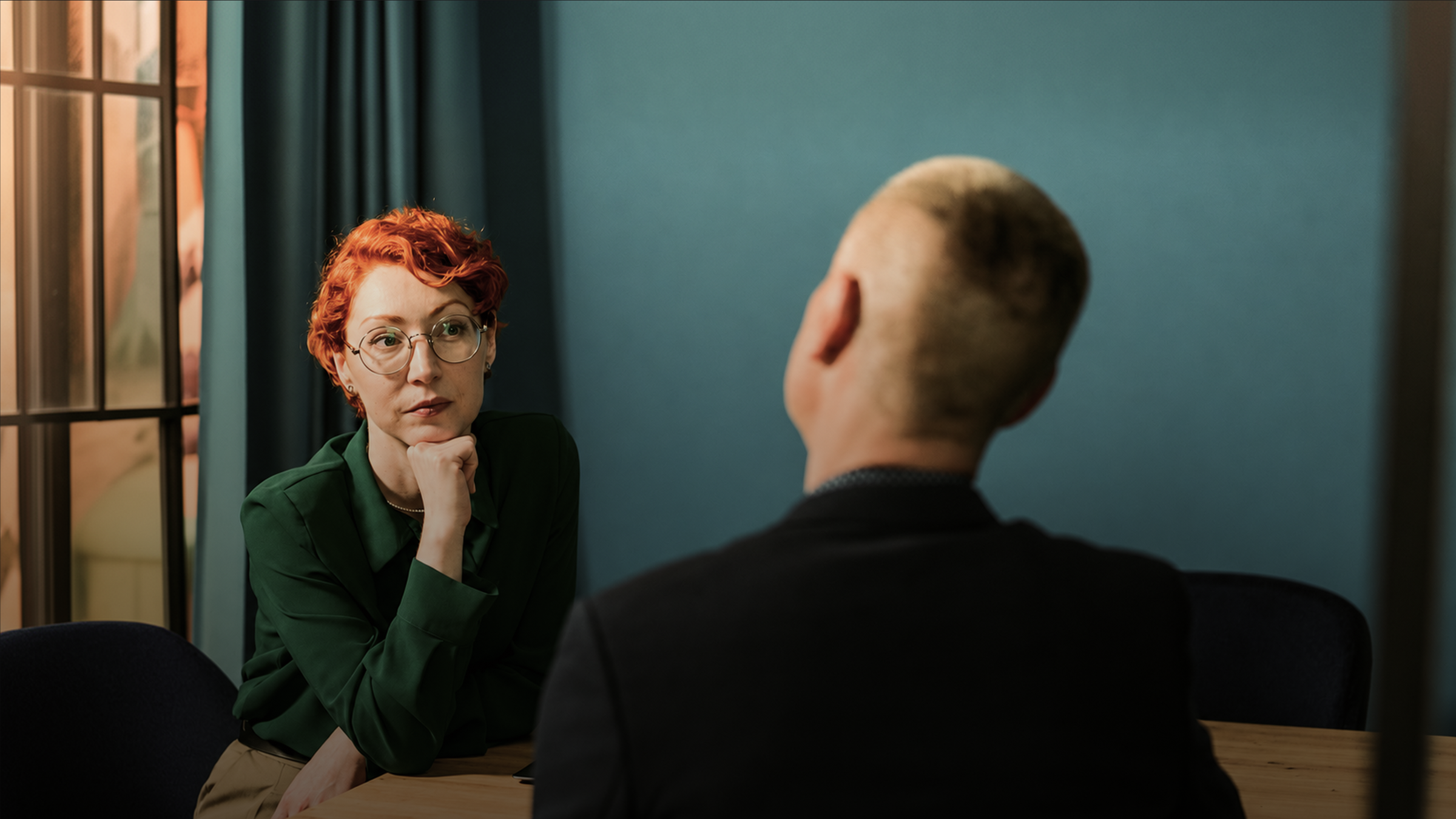 A red-haired woman in glasses listens intently to a person seen from behind across a table.