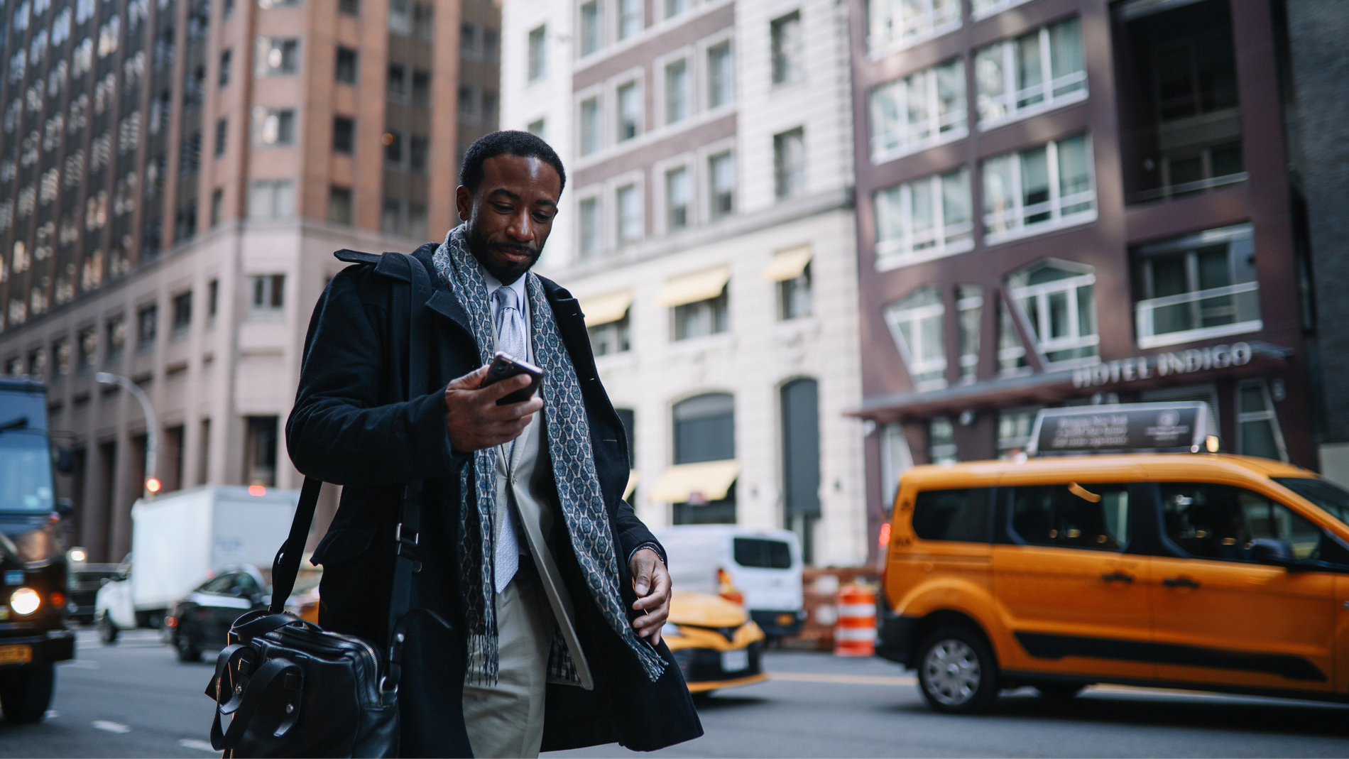 A Black man in a coat and scarf looks at his phone while walking on a busy city street.
