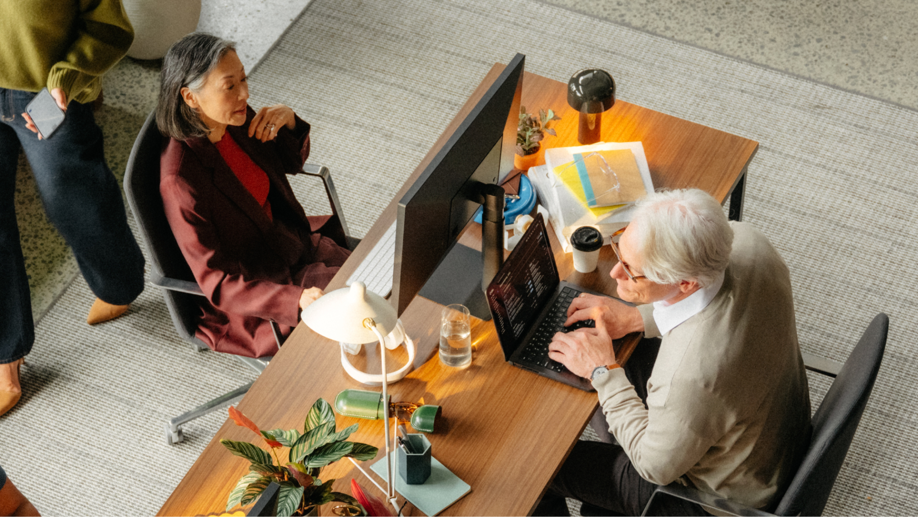 Overhead shot of two people at an office desk with computers, and a third person standing.
