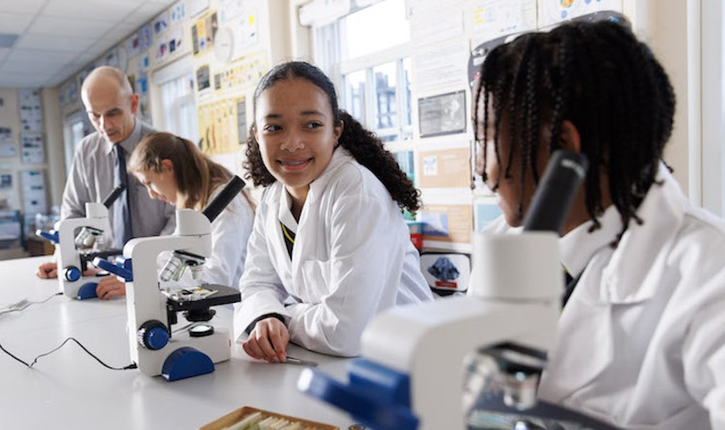 3 students wearing a lab coat and a teacher in a classroom with 3 microscopes on a table 