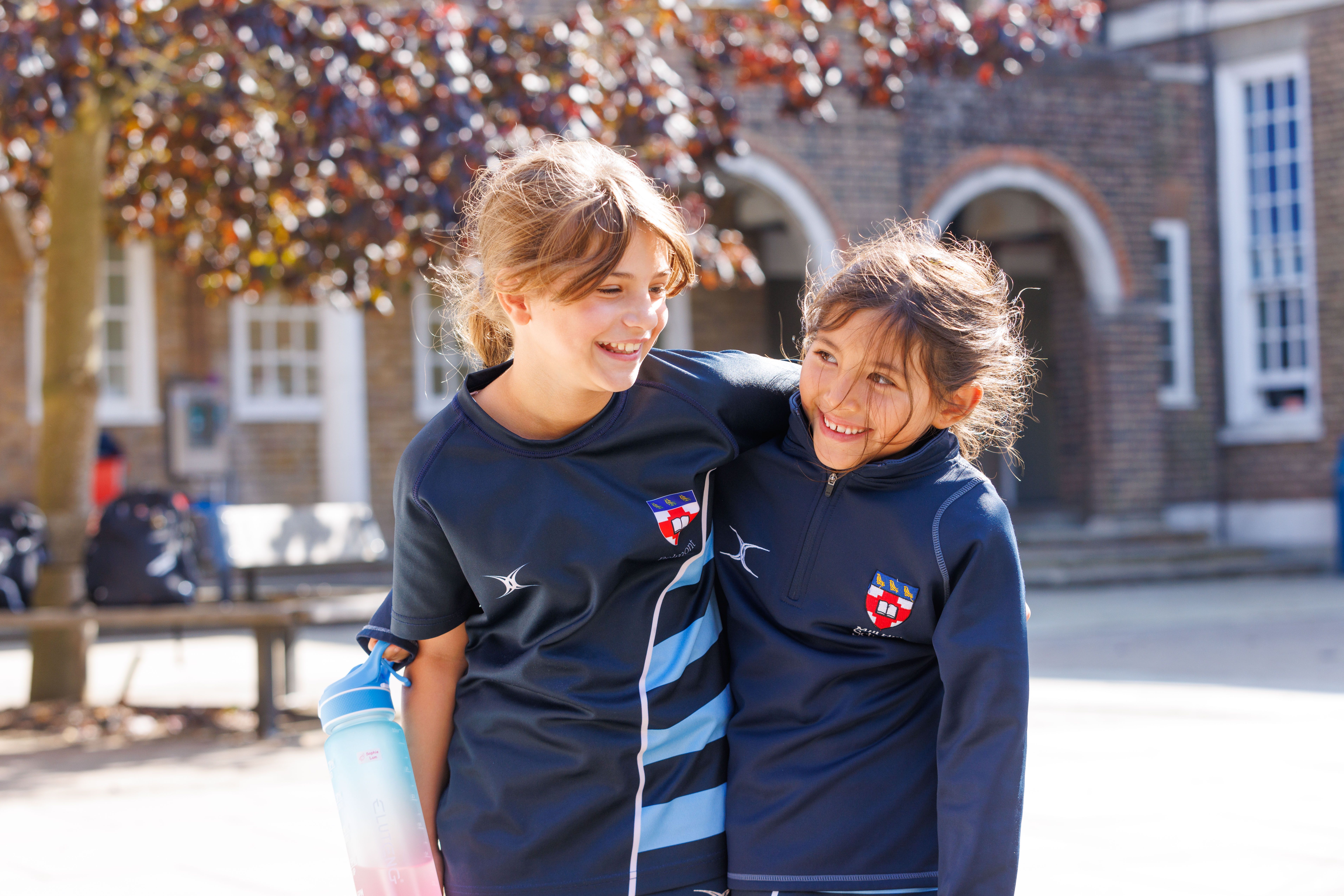 Two young girls outdoors smiling with their arms across each other from behind 
