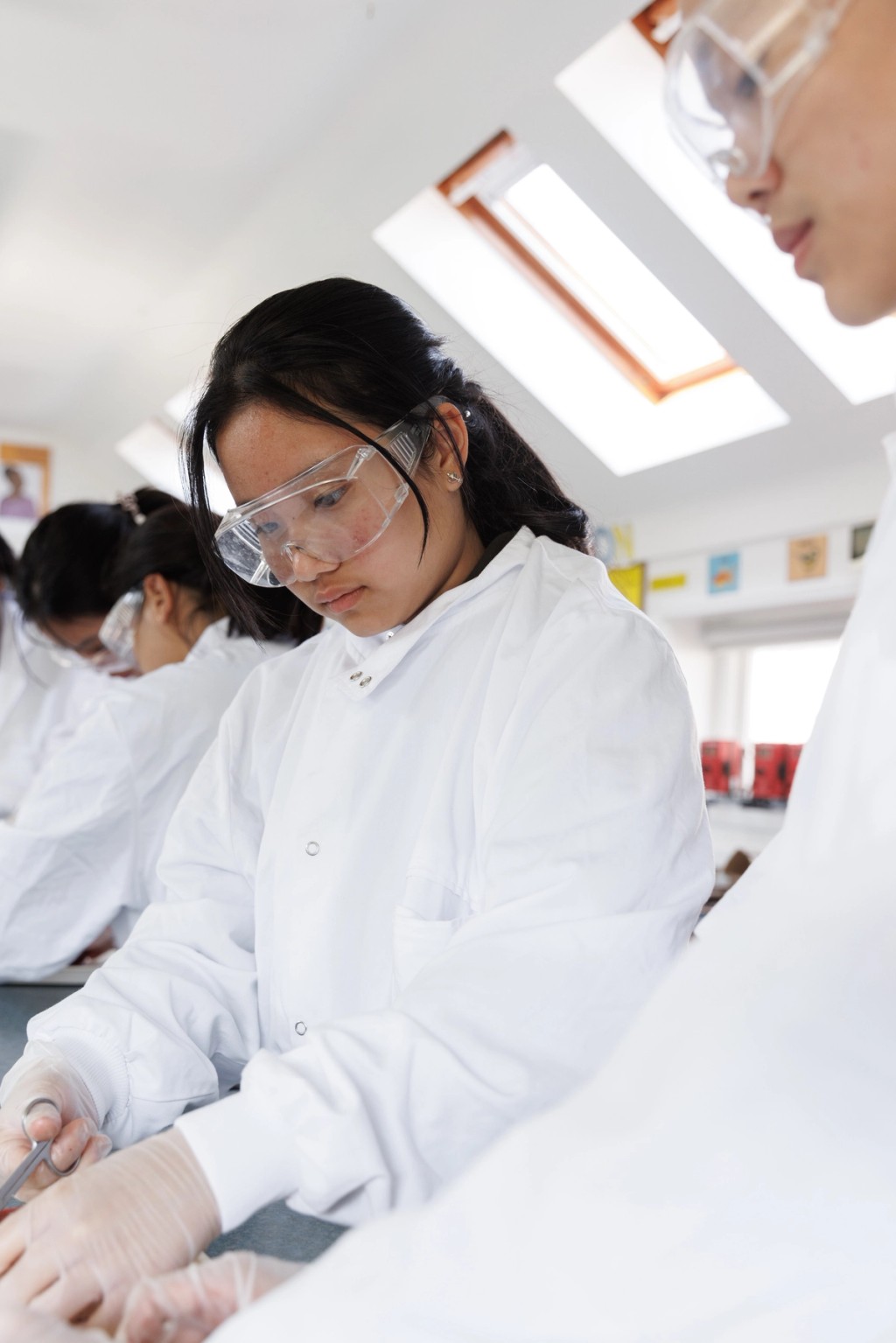 Image of students in a school science lab with a focus on a girl wearing goggles, looking down and holding a pair of scissors 