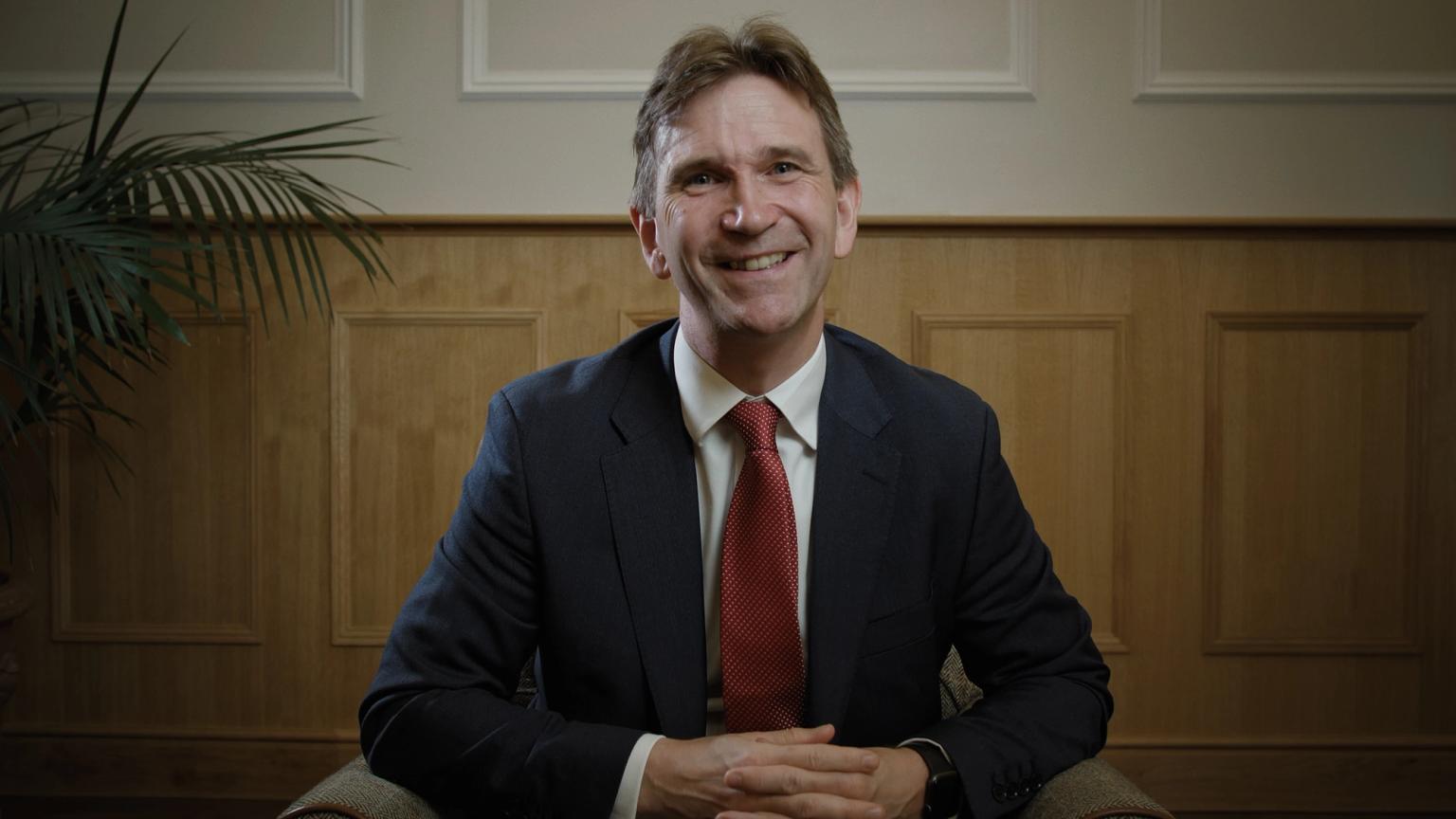 Image of a man, the CEO of Mill Hill Education Group, seated down, wearing a suit and smiling
