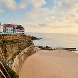 Buildings sit atop a rocky cliff overlooking a sandy beach and calm sea under a cloudy sky at sunset Portugal