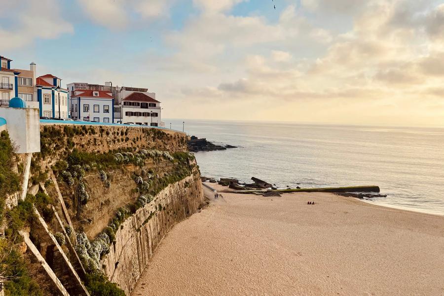 Buildings sit atop a rocky cliff overlooking a sandy beach and calm sea under a cloudy sky at sunset Portugal