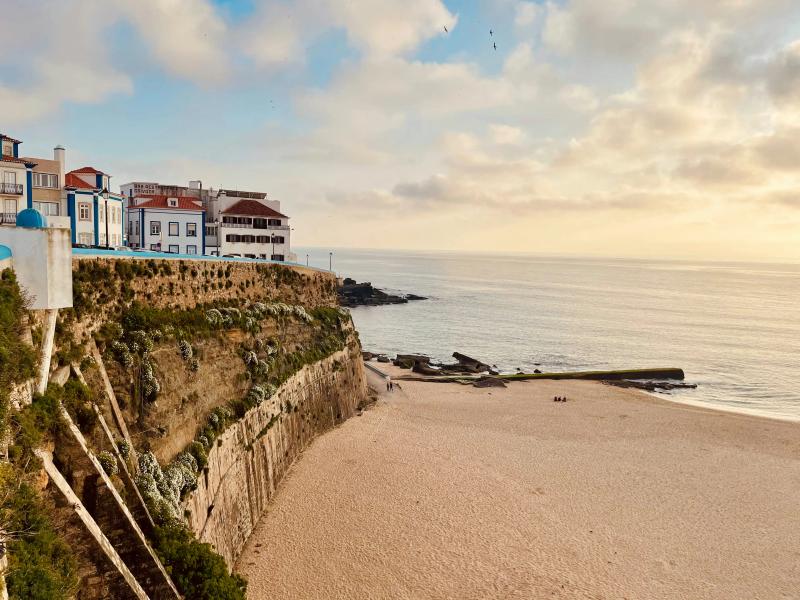 Buildings sit atop a rocky cliff overlooking a sandy beach and calm sea under a cloudy sky at sunset Portugal