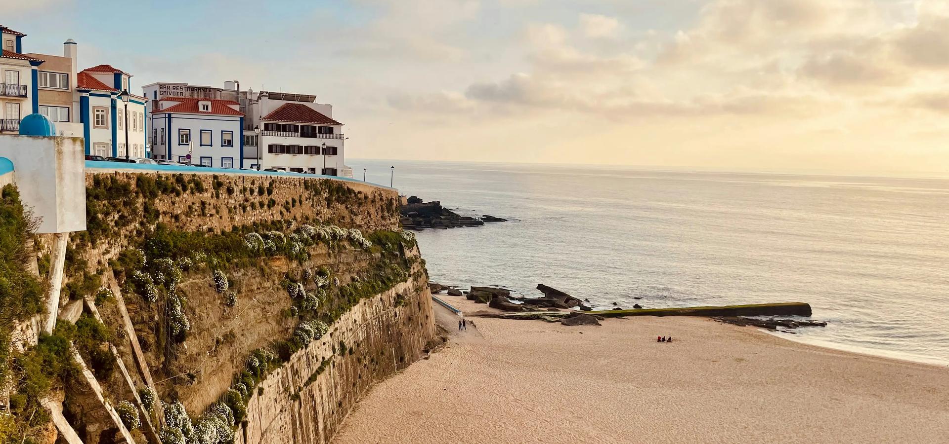 Buildings sit atop a rocky cliff overlooking a sandy beach and calm sea under a cloudy sky at sunset Portugal