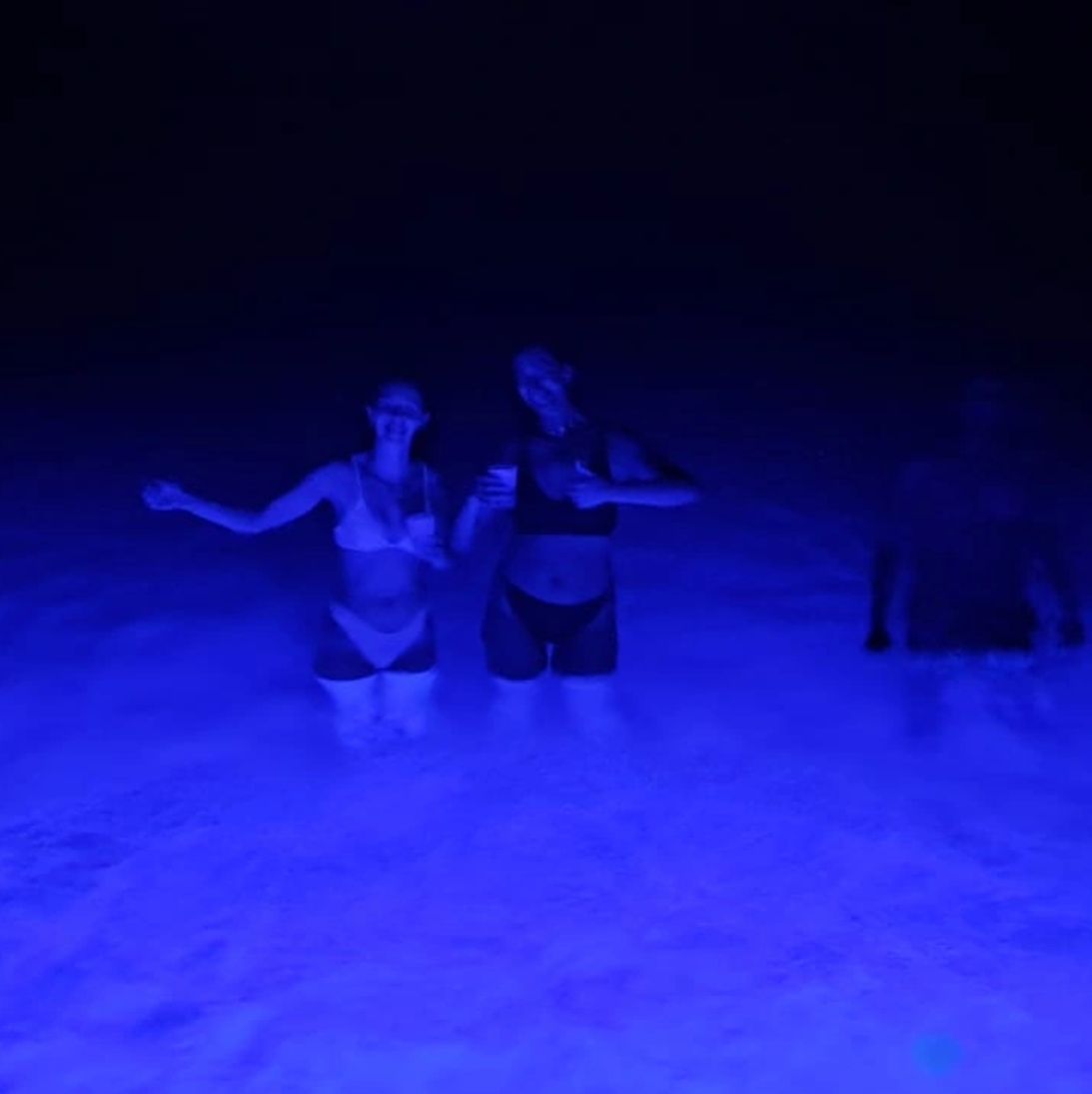 Two guests standing in the lagoon at night, the water around them glowing blue