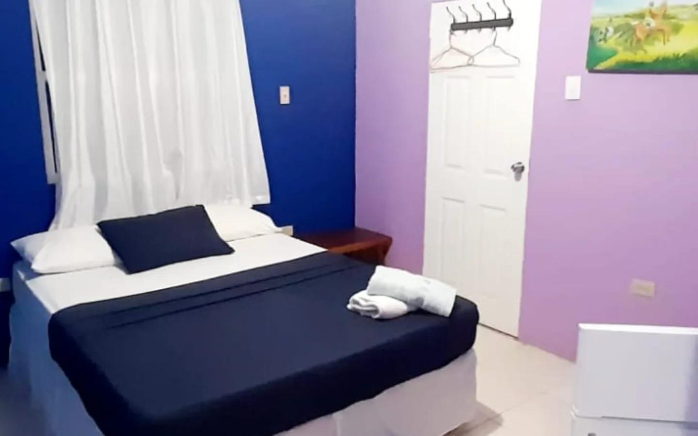 A queen bed made up in navy against a blue and lilac wall, with a coat rail on the door