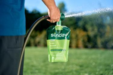 Person using a hose-end sprayer pouch of Sunday “Iron Boost” liquid lawn nutrients, spraying a lawn with water mist.