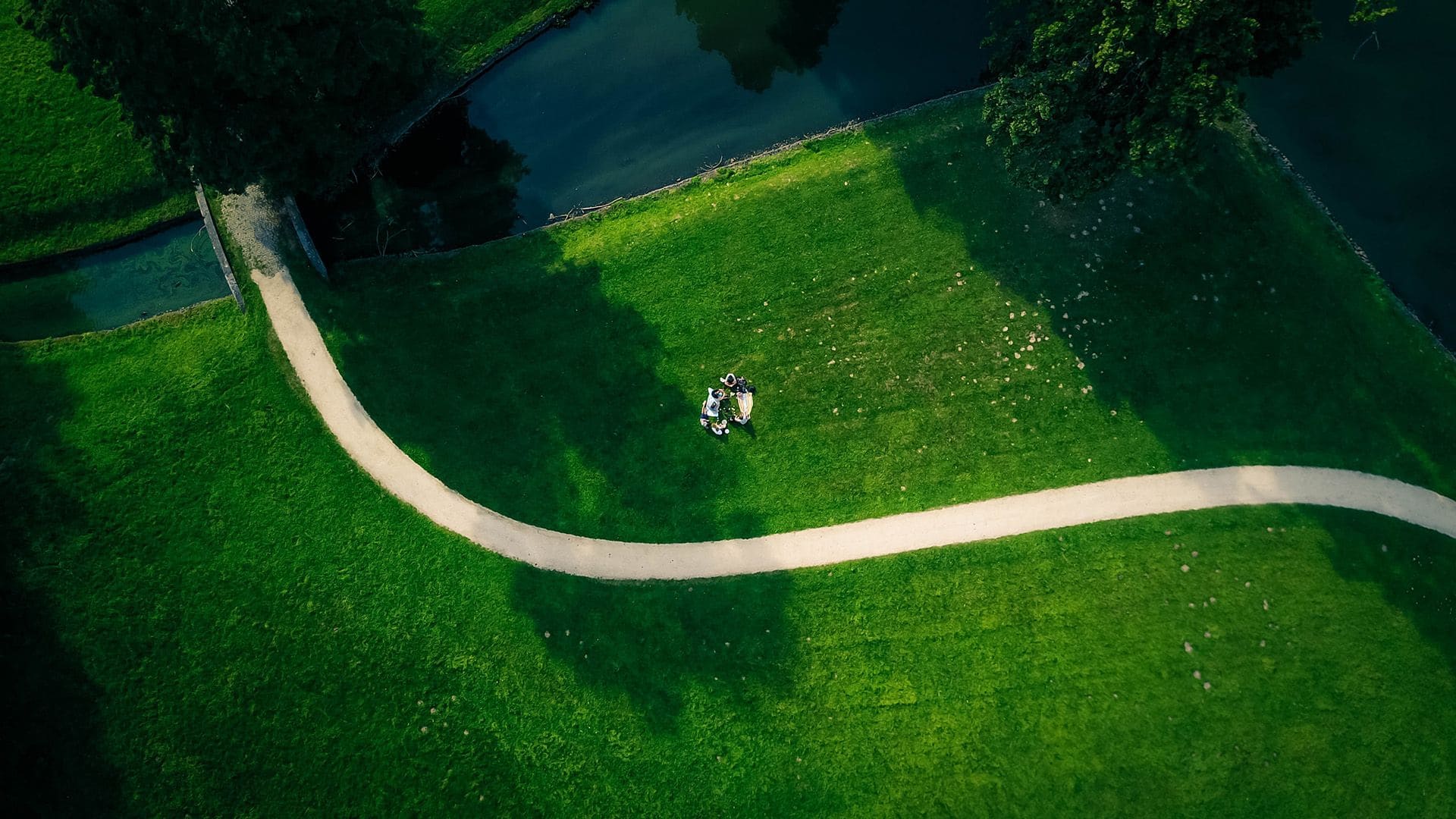Aerial view of a wide green lawn with a winding tan path and a small group of people gathered in a circle on the grass.