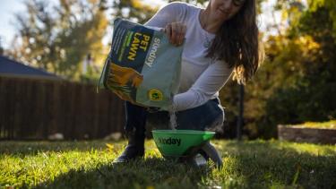 Woman pouring Sunday's Pet Lawn grass seed into handheld seed spreader over fall lawn for fall grass seeding.