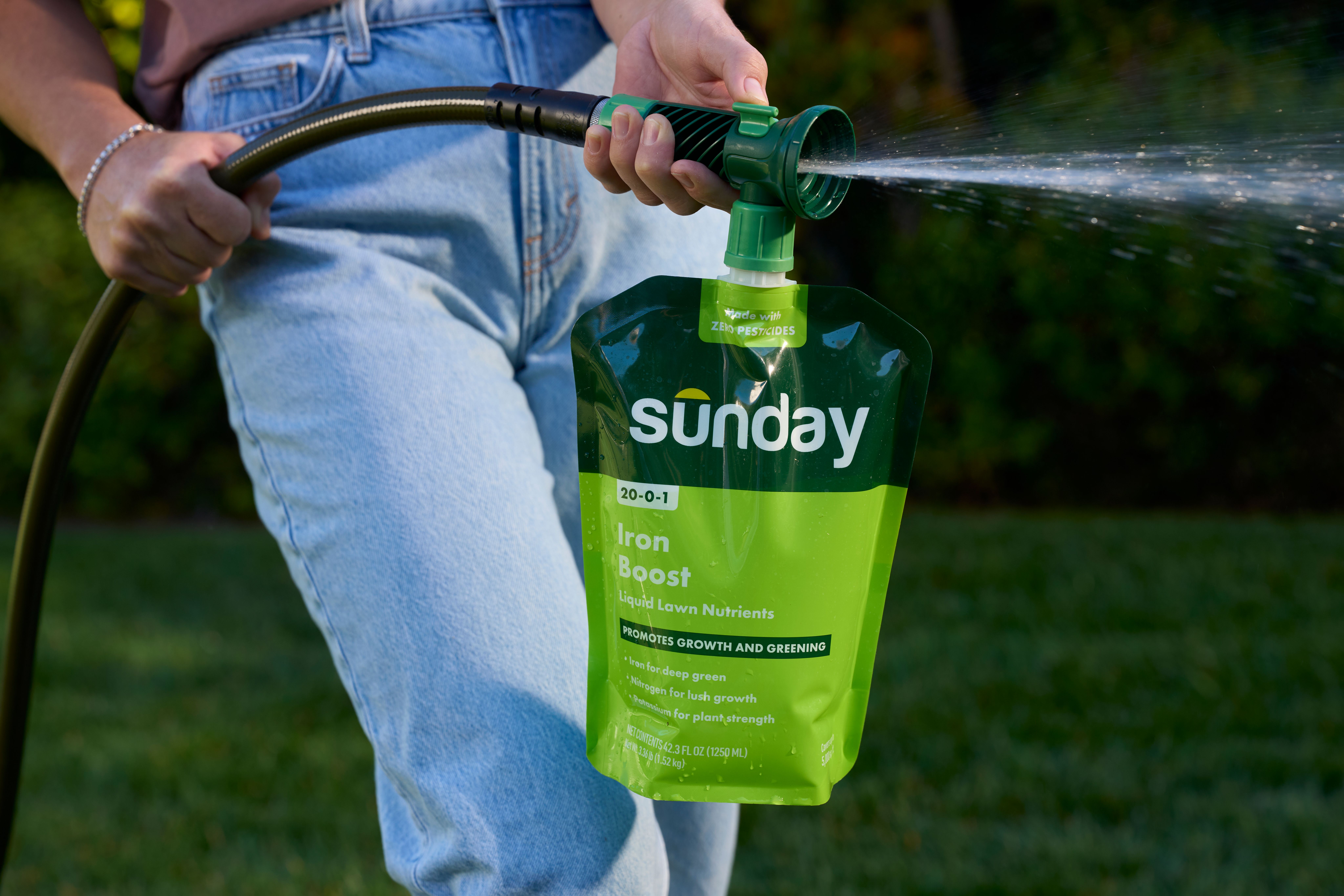 Close-up of a person using a hose-end sprayer pouch of Sunday “Iron Boost” liquid lawn nutrients, spraying a fine mist onto a lawn.