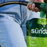 Close-up of a person using a hose-end sprayer pouch of Sunday “Iron Boost” liquid lawn nutrients, spraying a fine mist onto a lawn.
