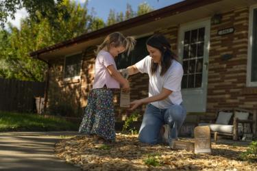 A woman helps a child fill a paper soil sample bag using a green-handled soil probe; Sunday kit boxes are on the mulch.